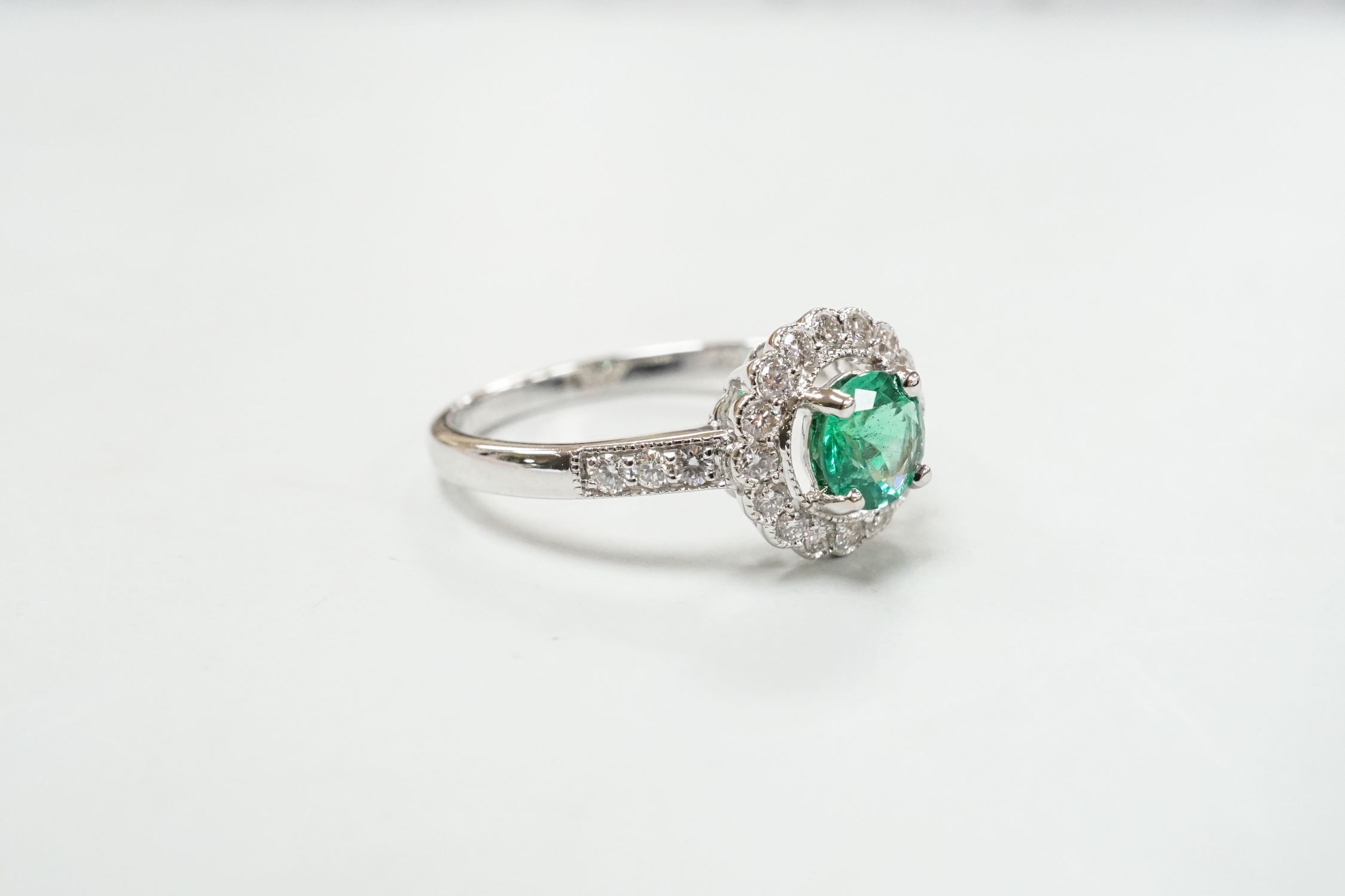 A modern 18ct white gold, emerald and diamond set circular cluster ring, with diamond set shoulders, size M, gross weight 2.7 grams.